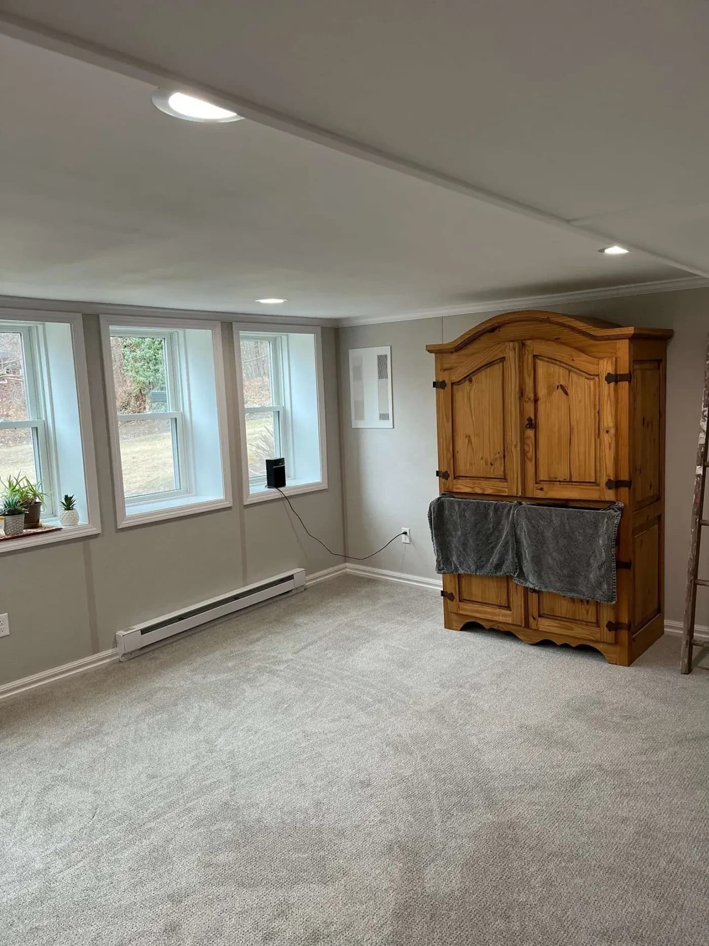 A bright basement room with plush carpet, recessed lighting, and three windows. A wooden cabinet with a towel hanging on it adds warmth. Peaceful ambiance.