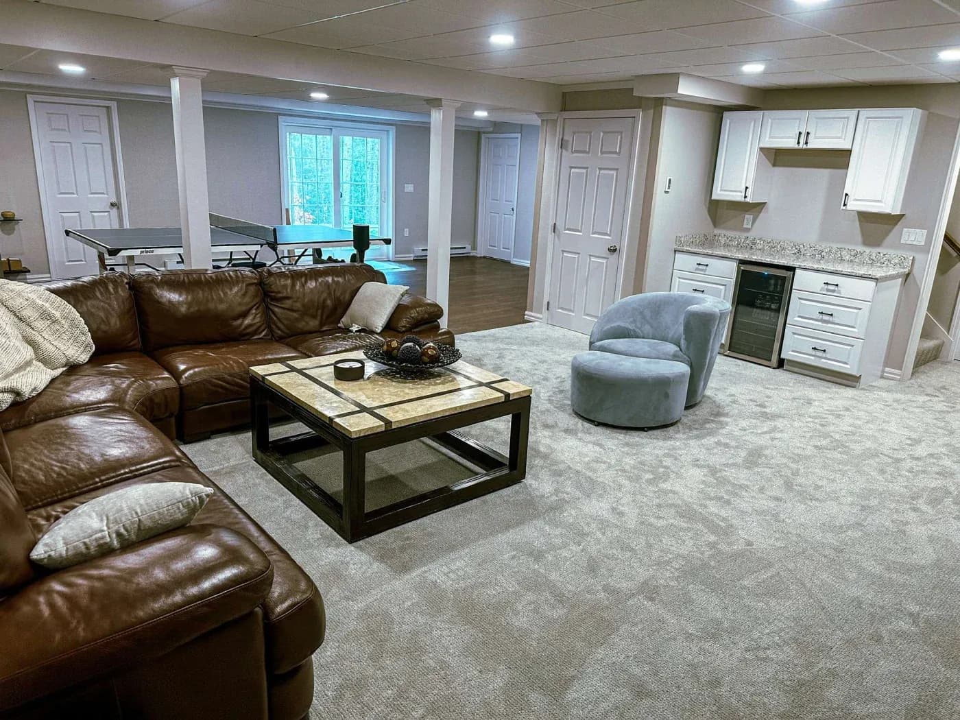 Spacious basement with brown leather sectional, beige carpet, and a tile coffee table. Features a kitchenette, a ping pong table, and cozy seating.