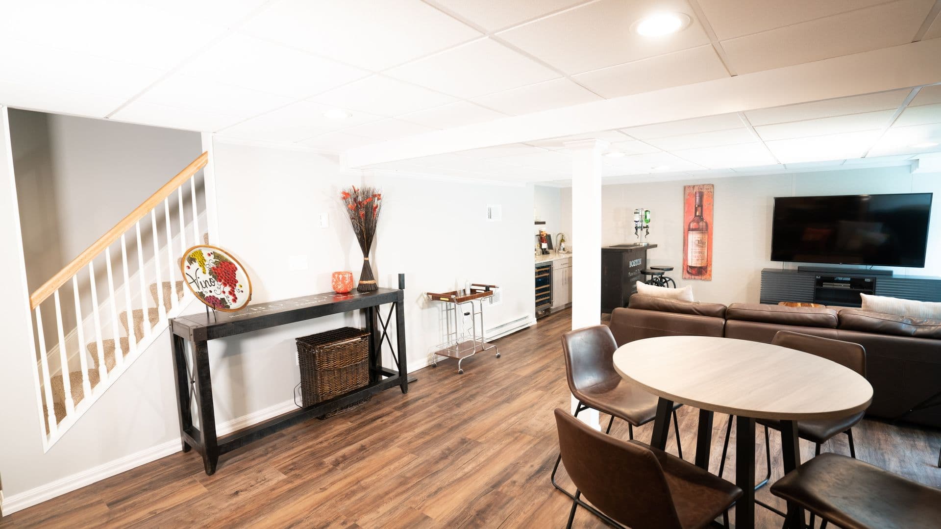 Modern basement with wood flooring, round table and four chairs, cozy sectional sofa, large TV on wall, console table with decor, and stairway.