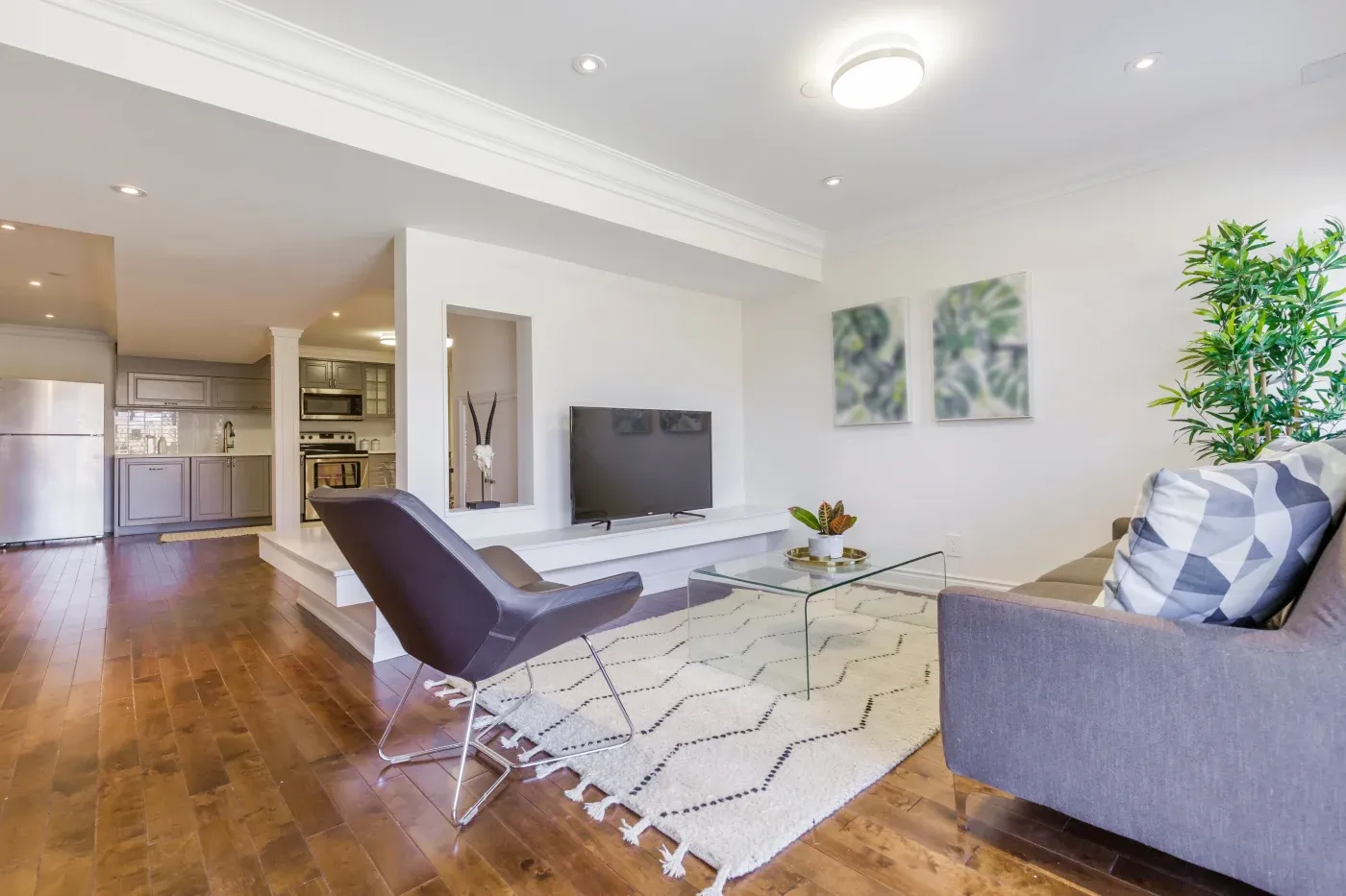 Modern living room with hardwood floors, featuring a gray sofa, black chair, glass coffee table, and wall-mounted TV. Open kitchen visible in the background.