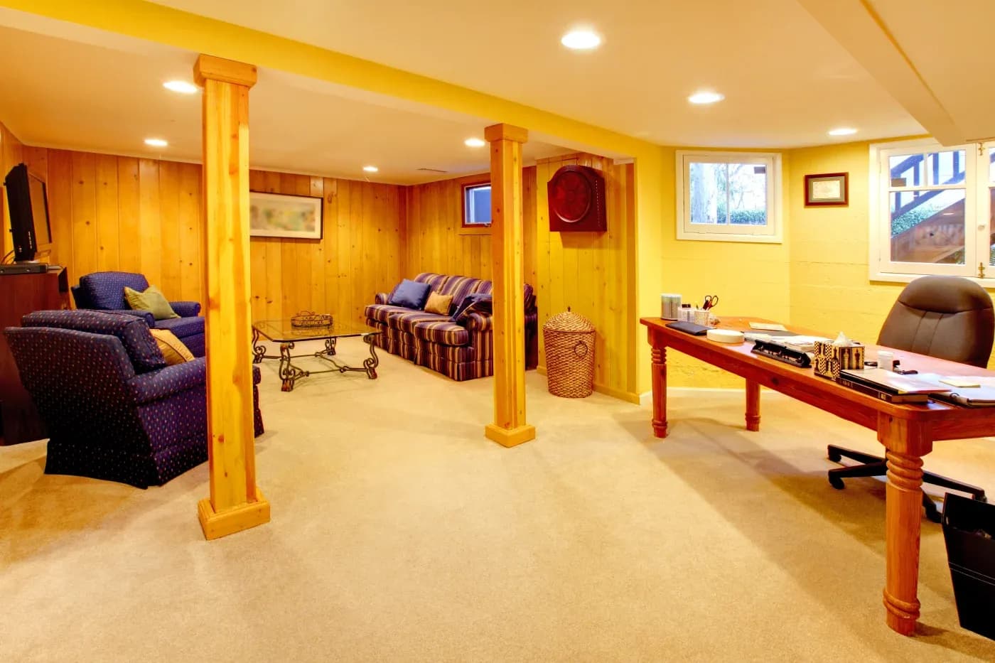 Warm, cozy basement with wood paneling and carpet, featuring a seating area with sofas, TV, and a wooden desk with chair, creating a relaxed atmosphere.