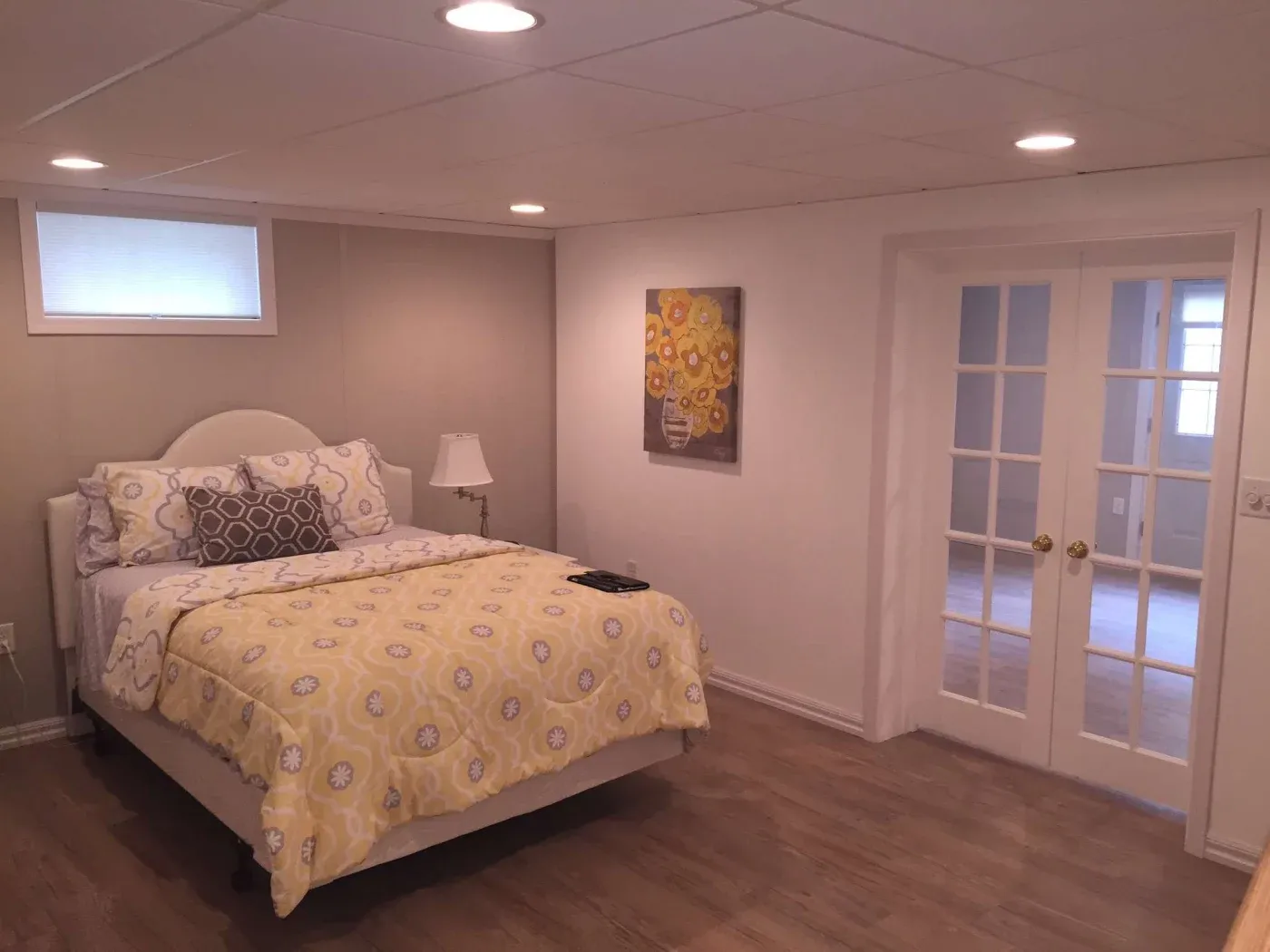 A cozy bedroom with warm lighting features a bed with yellow floral bedding, a bedside lamp, a floral painting, and French doors leading to another room.