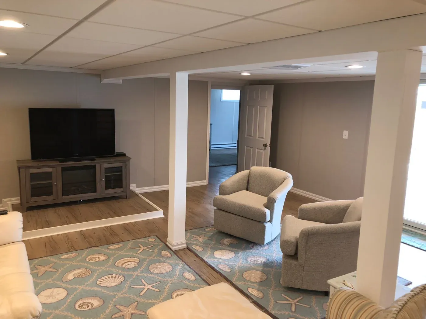 A cozy basement living room with beige walls features a TV on a wooden stand, two plush armchairs, and a sea-themed rug, creating a relaxed, inviting ambiance.