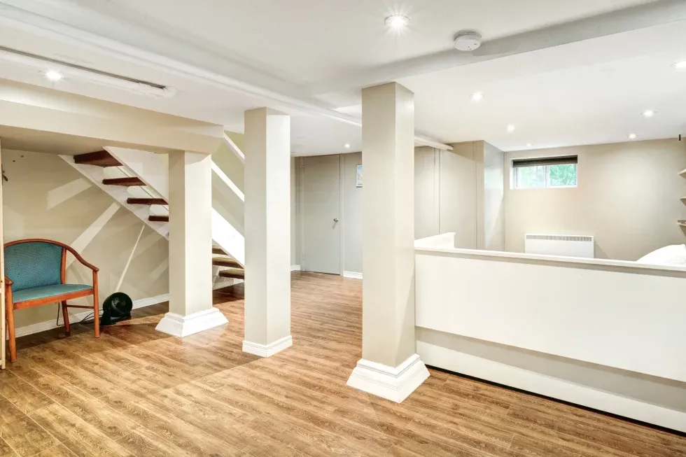 Spacious basement with light wood flooring, two support columns, and a minimalistic wooden staircase. A teal chair sits in the corner, creating a cozy atmosphere.