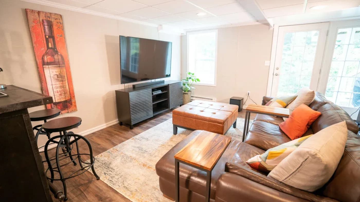 Cozy living room with a brown leather sofa, colorful pillows, and a large wall-mounted TV. A wine-themed artwork, wooden floor, and soft lighting create a warm ambiance.