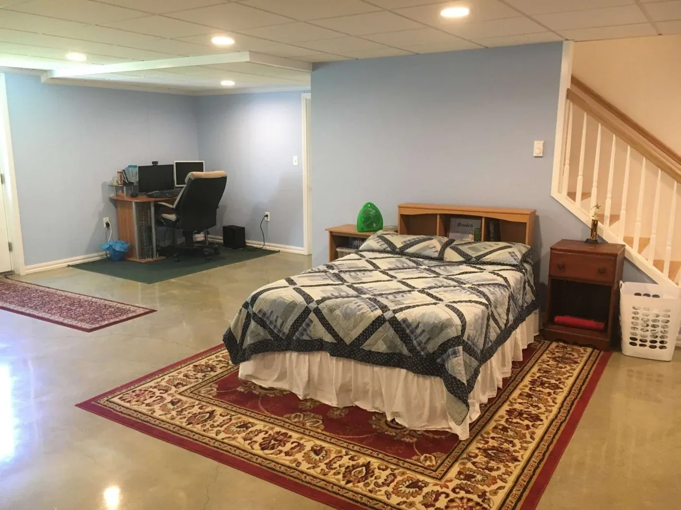 A basement room with pale blue walls, featuring a bed with a patterned quilt, a desk with a computer setup, and a staircase. The floor is polished with red and beige rugs.