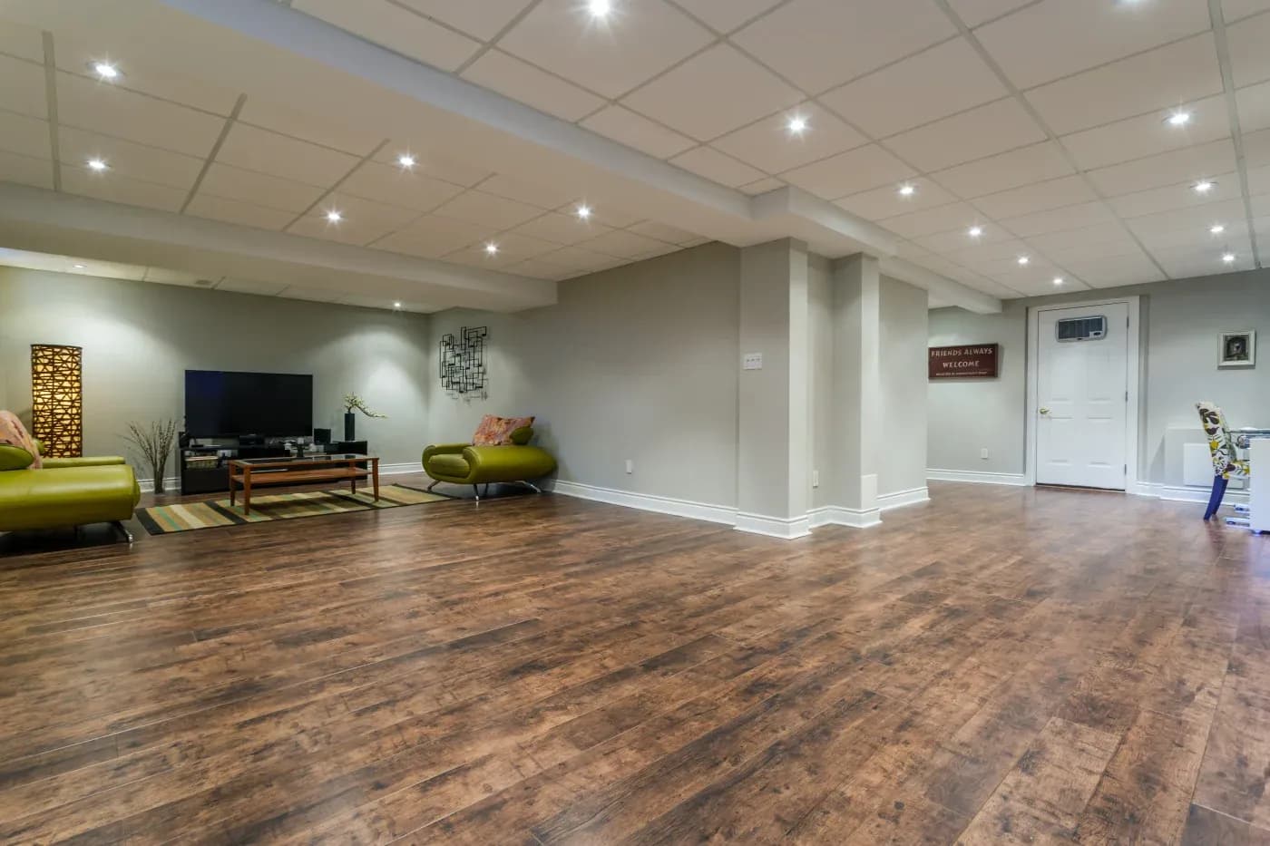 Spacious basement with polished wooden flooring, cozy green sofas, a flat-screen TV on a wooden stand, and soft recessed lighting. Modern and welcoming.