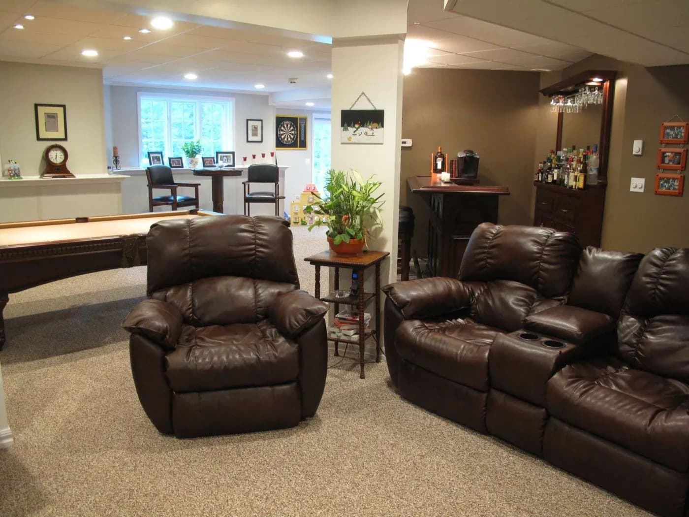 Cozy basement living area with brown leather recliners, a shuffleboard table, and a bar. Brightly lit with plants and game elements like a dartboard.