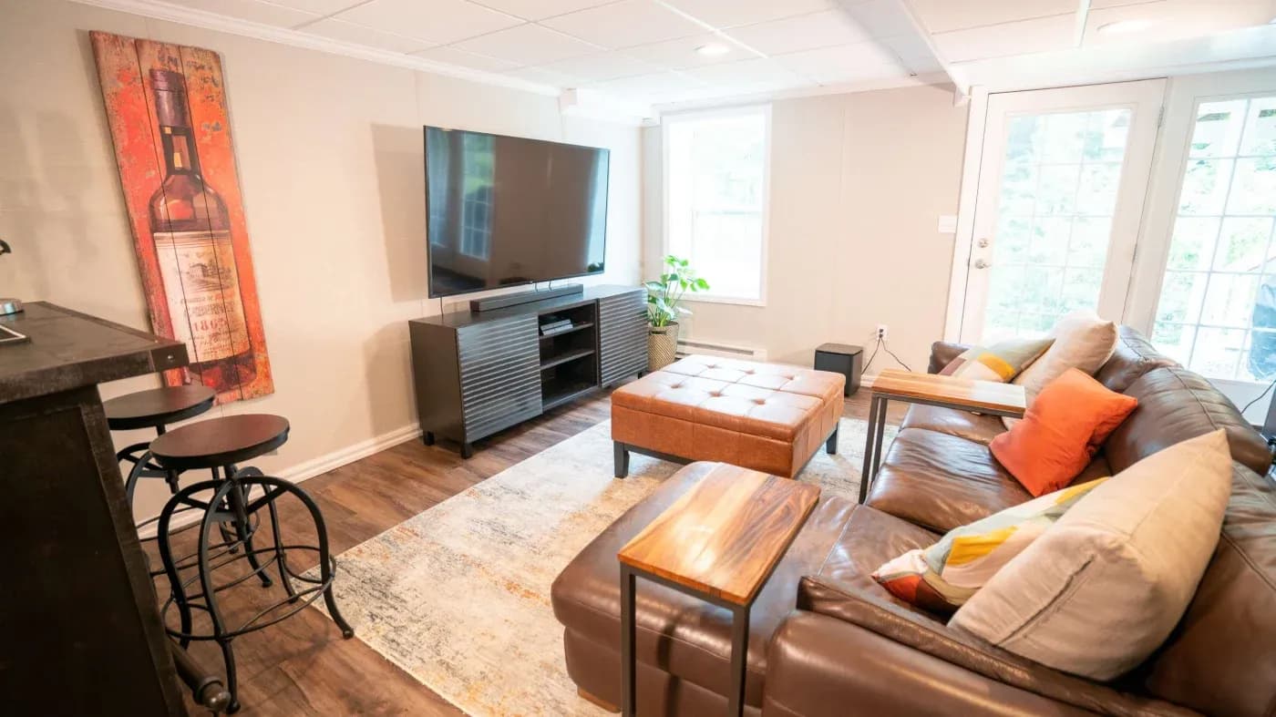 A cozy living room with a leather sectional sofa, colorful pillows, and a wooden coffee table. Large TV on a console, wall art of a wine bottle, bar stools, and bright natural light from a window and glass doors. Warm, inviting atmosphere.