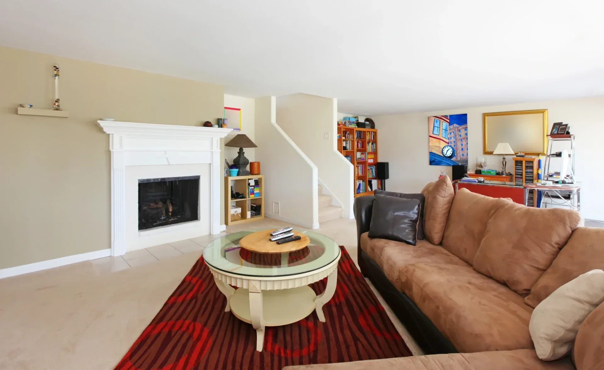 A cozy living room with a beige sofa, decorative cushions, and a round glass coffee table on a red patterned rug. A white fireplace and colorful wall art add warmth, while bookshelves and a staircase create a welcoming, lived-in atmosphere.