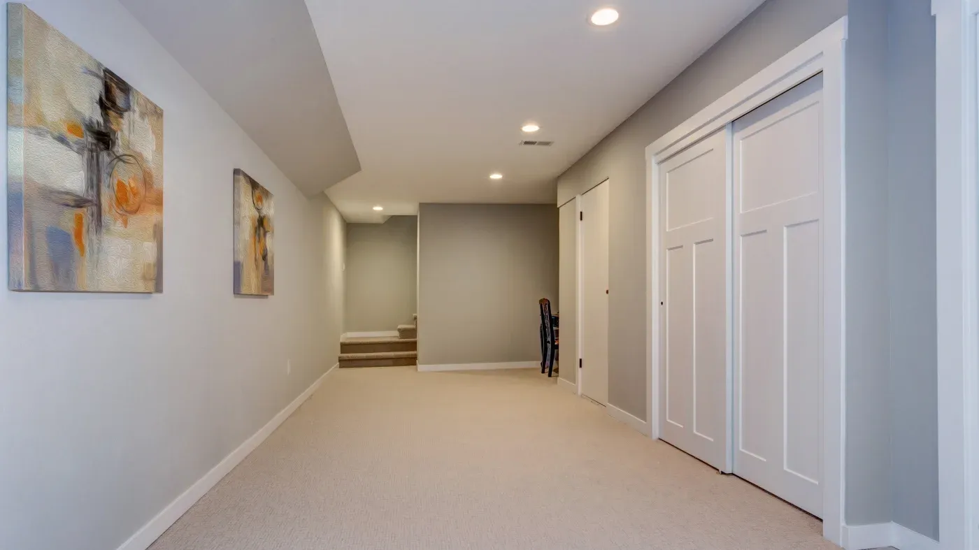 A spacious, empty hallway with gray walls, beige carpet, and two abstract paintings. Recessed lighting adds a warm tone. White sliding doors are seen on the right.