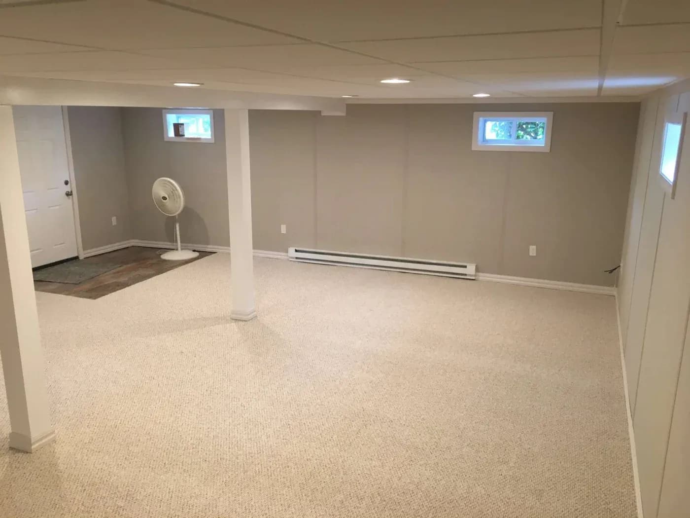 Spacious, empty basement room with beige carpet and neutral walls. A white fan stands near a door and small windows provide natural light.