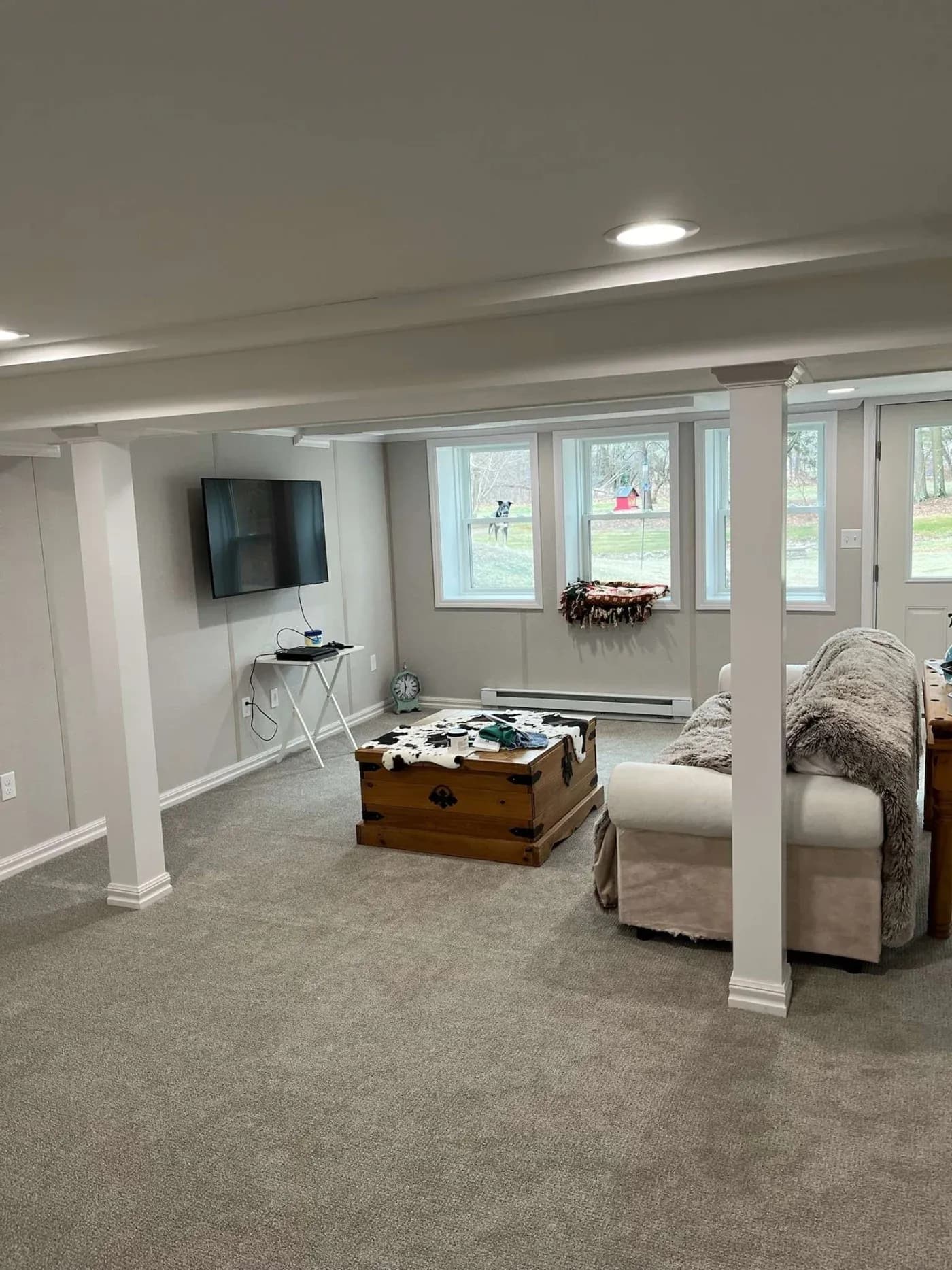 Basement room with beige carpet, featuring a sofa and wooden trunk as a table. A flat-screen TV is mounted on the wall. Windows show a grassy yard.