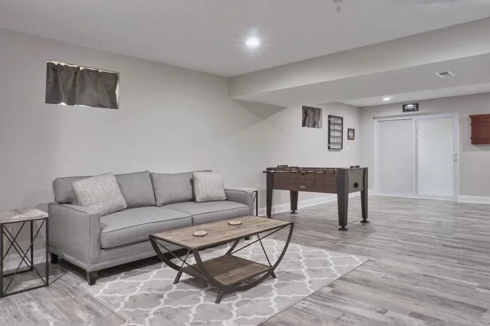 A modern basement with a relaxed atmosphere. Features a gray sofa, a patterned rug, and a foosball table on wood flooring. Walls are neutral-toned.
