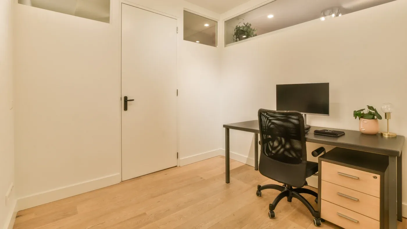 Modern office space with a minimalist design features a black desk, chair, computer, and drawer unit. Warm lighting and a potted plant add a cozy touch.