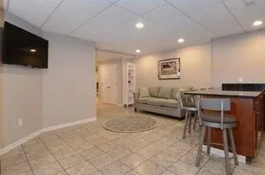 A cozy basement room with a beige tiled floor, gray walls, and recessed lighting. Features a wall-mounted TV, a gray sofa, a round rug, and a bar table with stools.