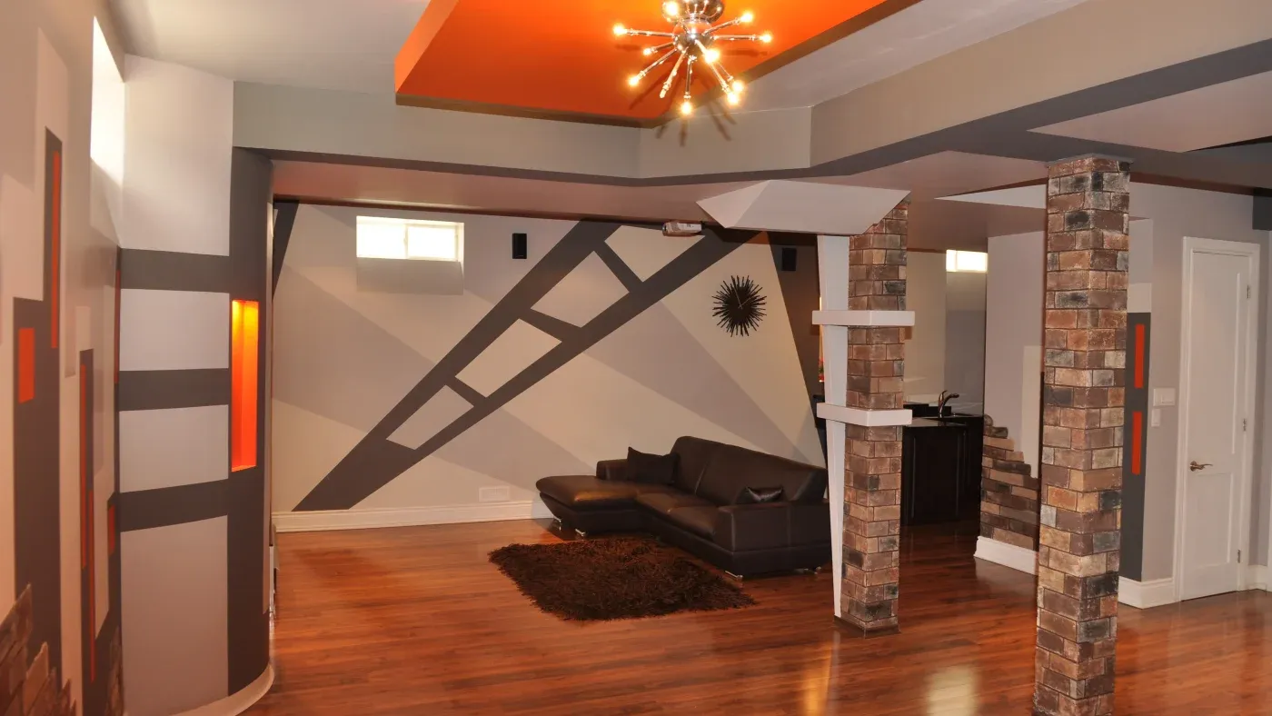 Modern living space with geometric wall designs and orange accents. A dark leather sofa, rug, exposed brick columns, and a starburst light fixture enhance the room's contemporary feel.