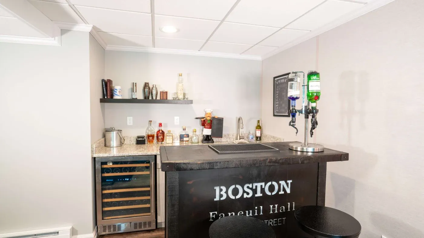 A home bar with "Boston Faneuil Hall" sign features a countertop with liquor bottles, a wine fridge, and two black stools, creating a cozy atmosphere.