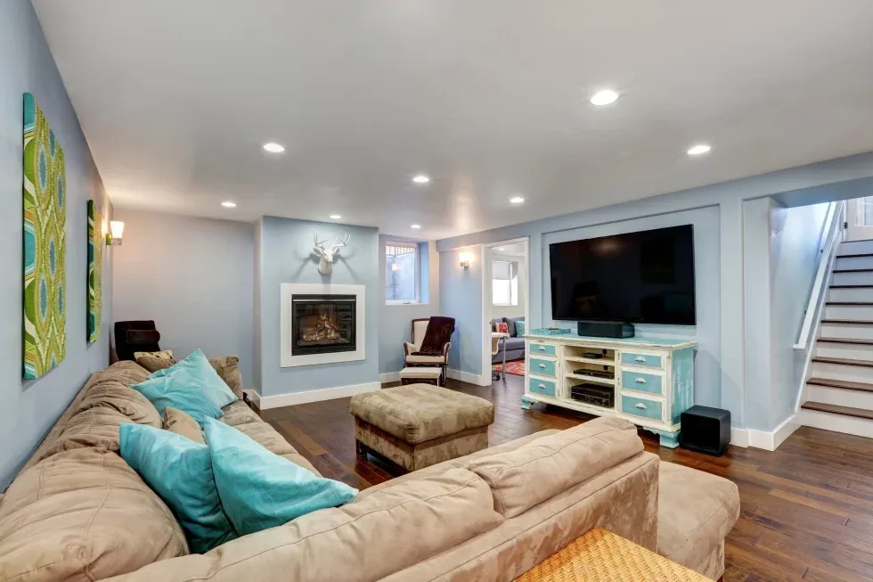 Spacious basement living room with beige sectional sofa, teal pillows, wall-mounted fireplace, large TV on a rustic cabinet, and modern decor. Cozy ambiance.