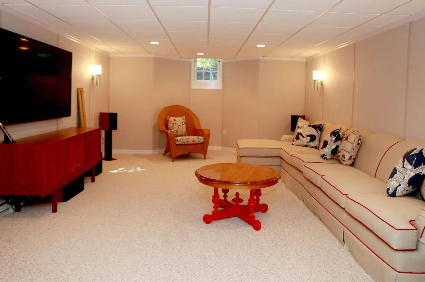 A cozy basement living room with a large beige sectional sofa adorned with patterned cushions, a red round coffee table, and a flat-screen TV on a wooden console. There's a wicker chair and a small window providing natural light, creating a warm and inviting atmosphere.
