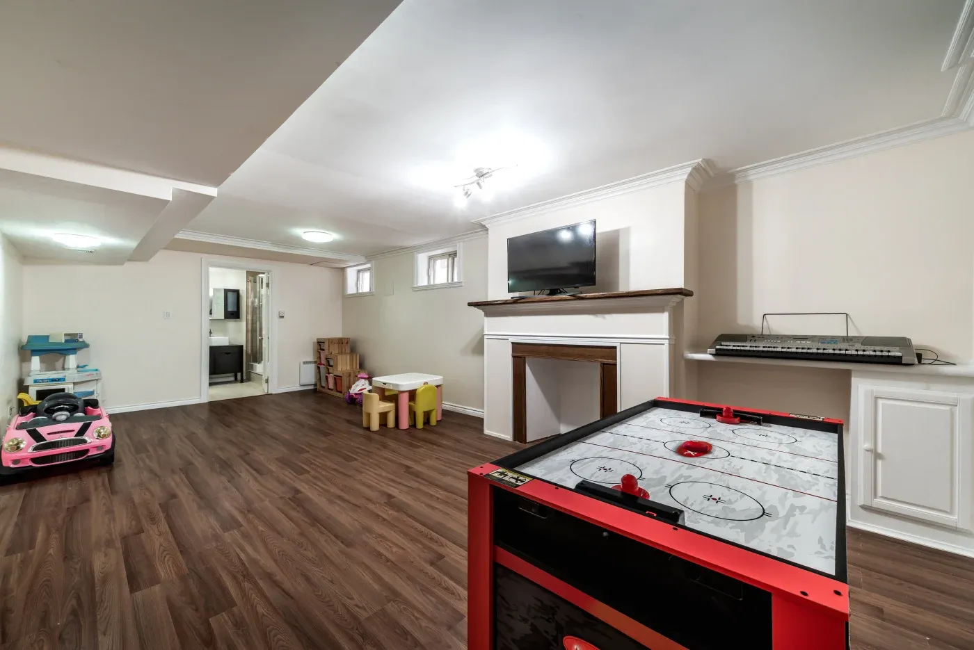 A spacious basement playroom with wooden flooring features a small pink toy car, air hockey table, TV above a fireplace, keyboard, and kids' table set.