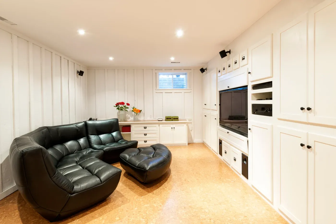 A cozy basement living area with a glossy black sectional sofa and ottoman on wooden floors. White cabinets line the walls, bright lighting overhead.