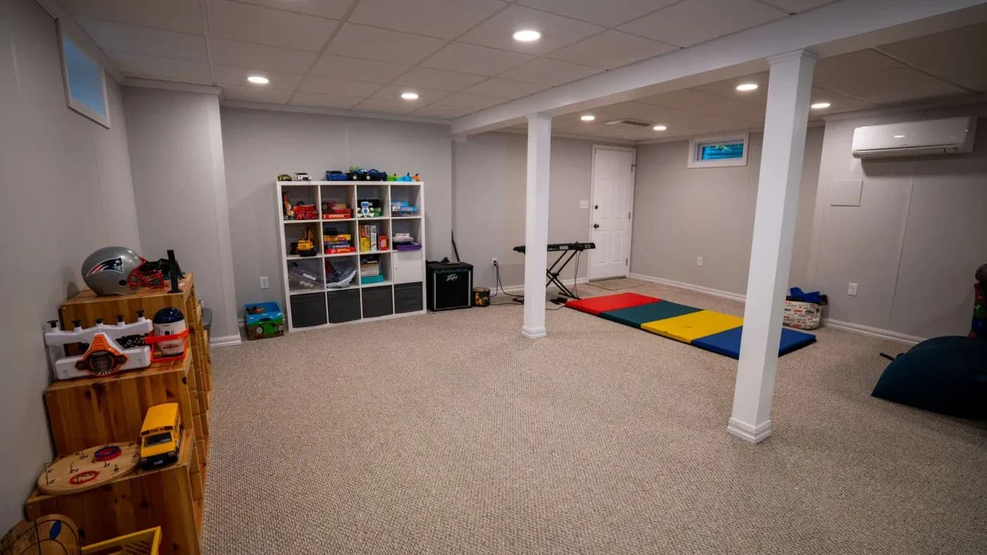 A spacious, carpeted playroom with a white ceiling and walls features colorful toy shelves, a small keyboard, and gym mats, creating an inviting, playful atmosphere.