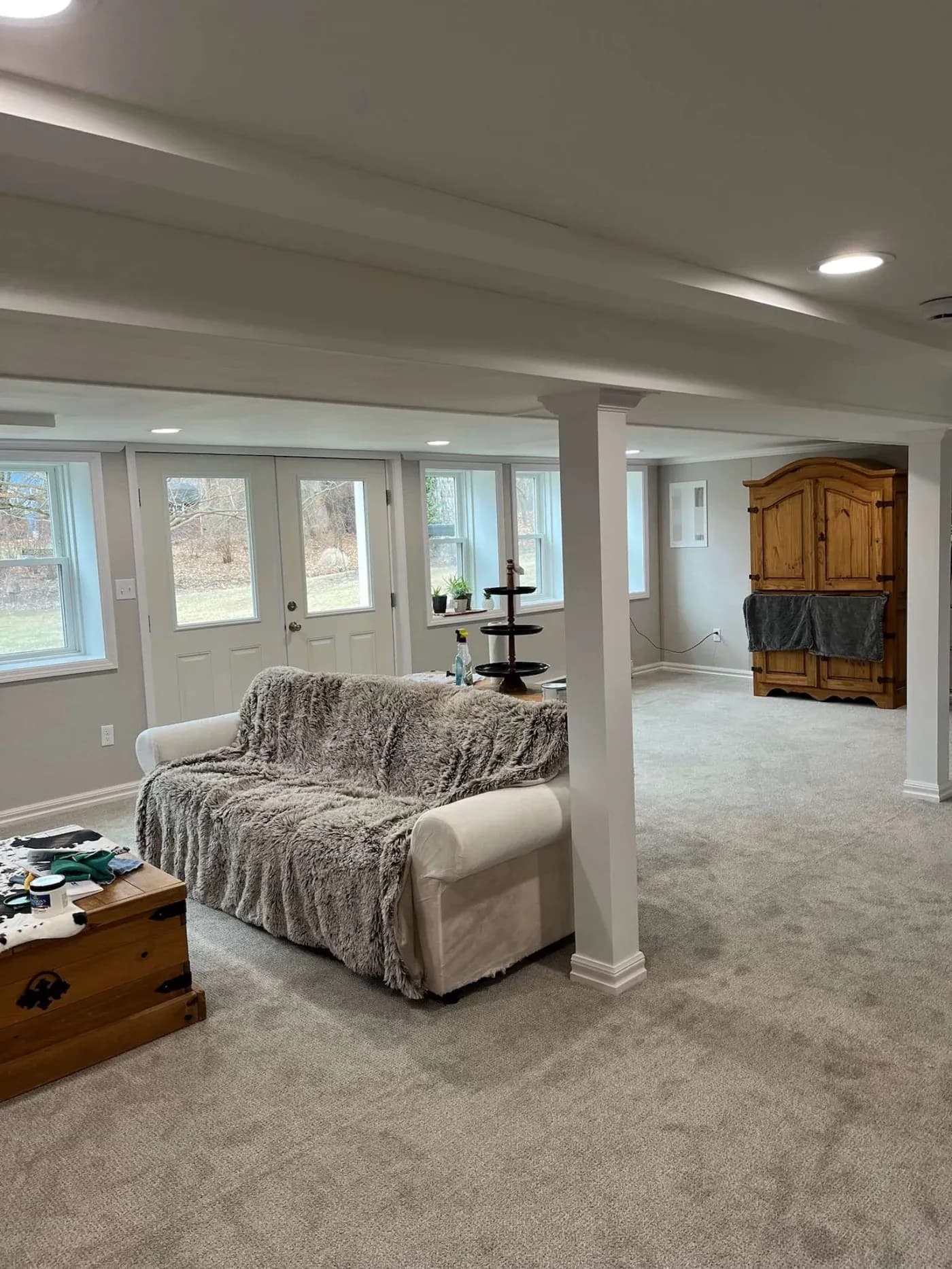 Spacious basement living area with neutral tones. Features a white sofa with a gray throw, wooden armoire, and natural light from multiple windows. Cozy atmosphere.