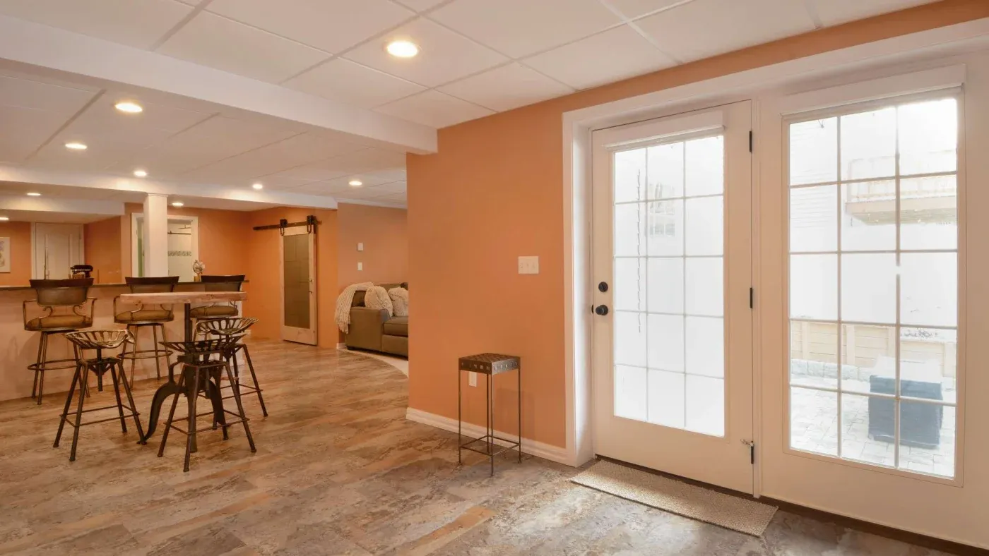 Spacious basement with warm orange walls, white ceiling, and wood-like flooring. Features barstool seating, a couch, and glass doors leading outside. Cozy ambiance.
