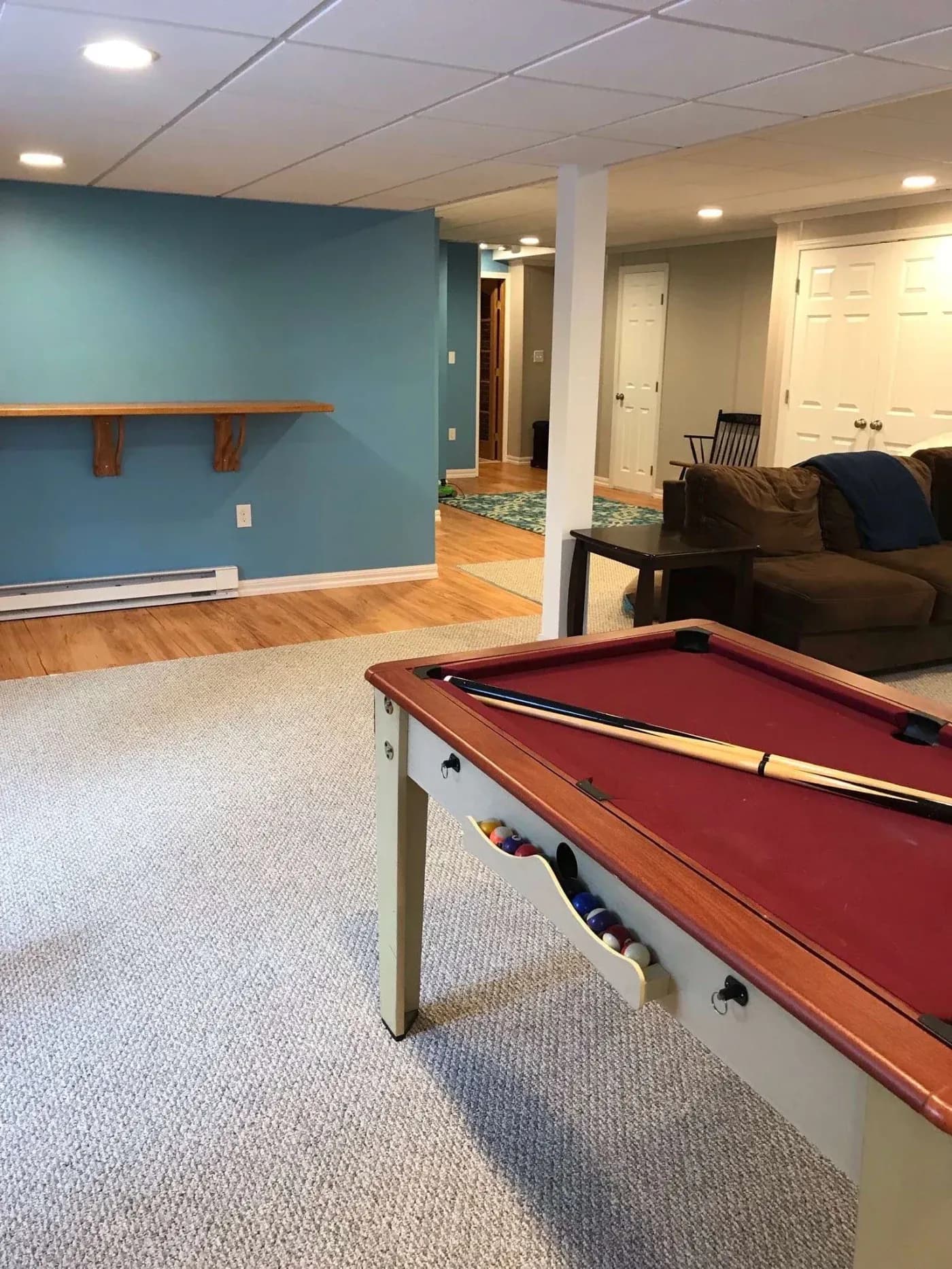 A cozy basement features a pool table with a red felt top and cue stick. Blue painted wall, shelf, brown sofa, and warm wood flooring accent the space.