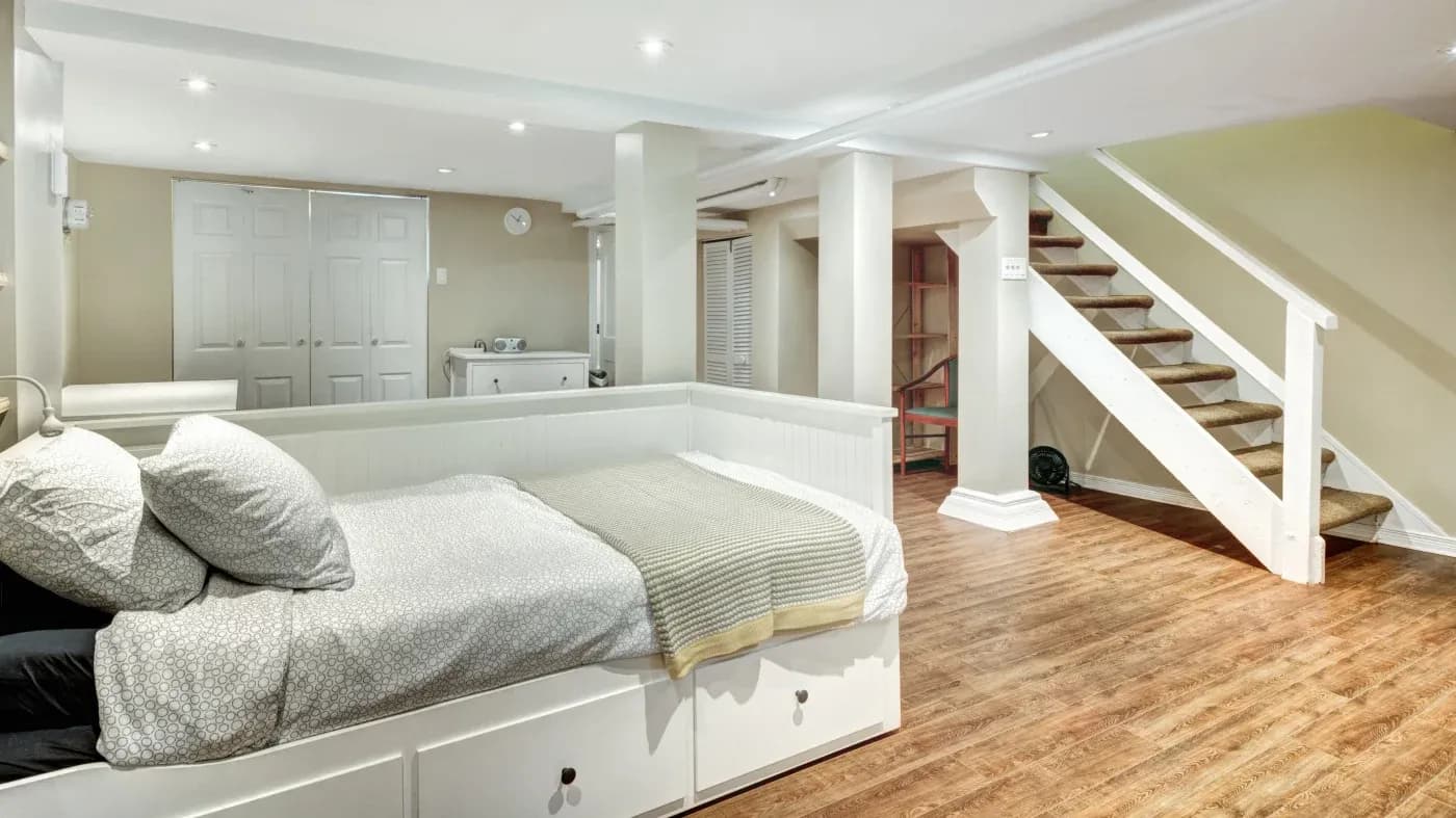 A cozy, well-lit basement bedroom features a daybed with patterned sheets and a green throw. Wooden floors, a staircase, and closet doors add warmth.