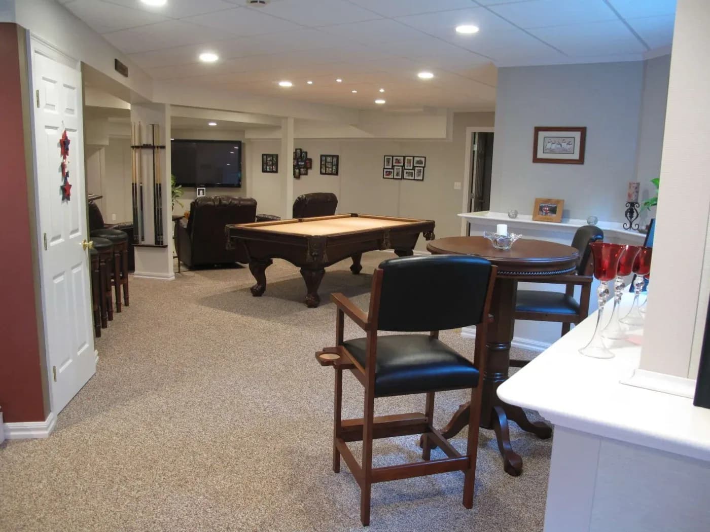 Spacious room with a pool table, bar stools, and a round table, featuring warm lighting. Comfortable seating and wall decor create a cozy, inviting atmosphere.