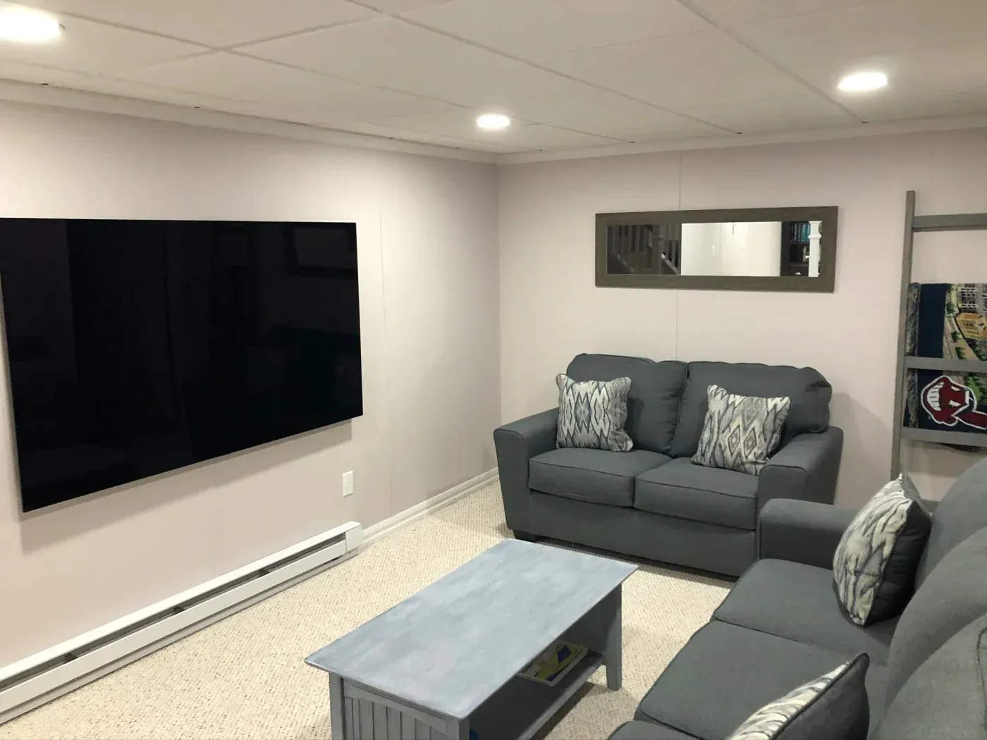 A cozy living room with light walls features a large flat-screen TV, a gray sofa set with patterned cushions, a blue-gray coffee table, and a wall mirror.