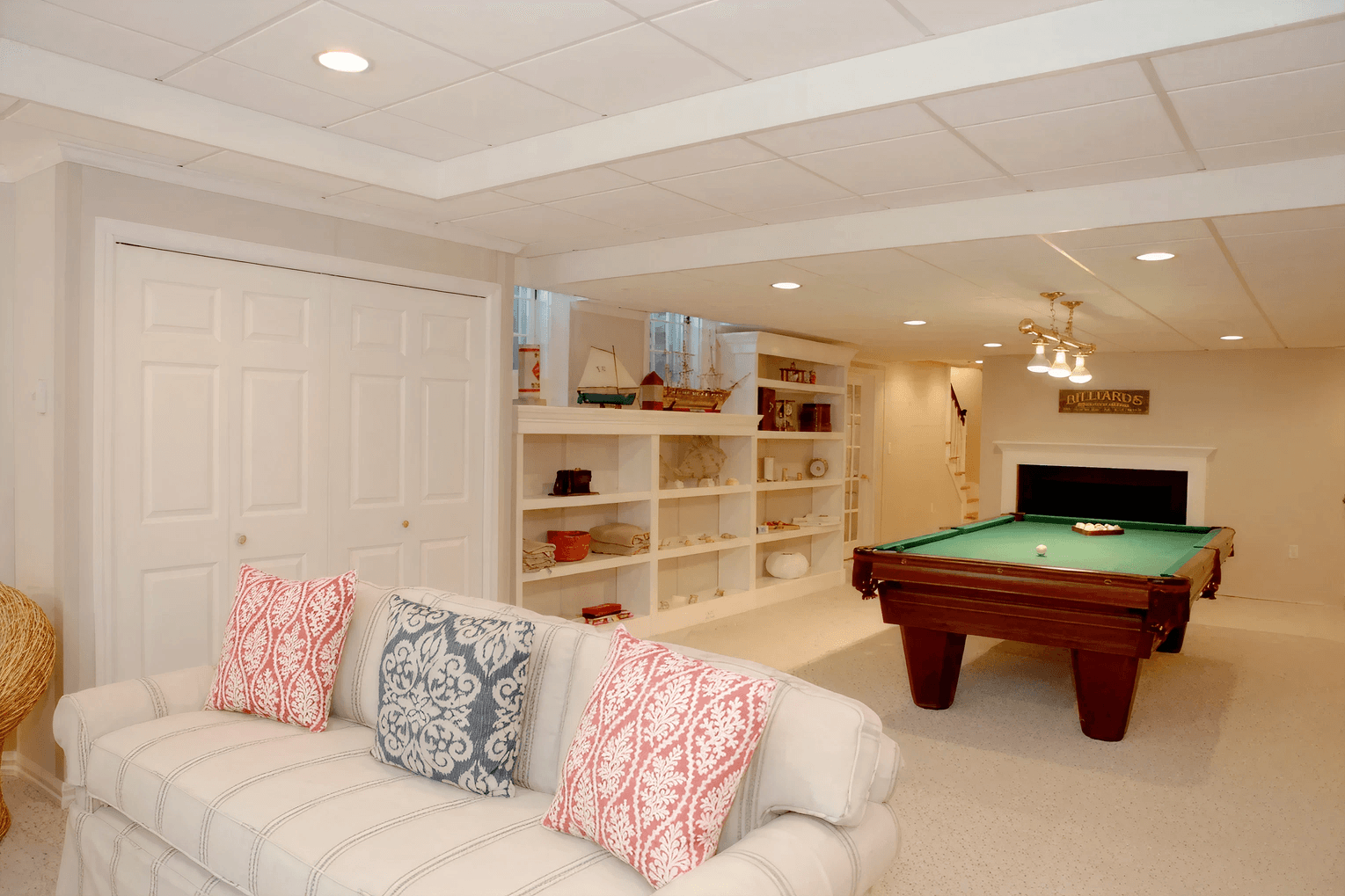 Bright basement lounge with a plaid sofa and patterned pillows, a pool table, and built-in shelves. Warm lighting creates a cozy atmosphere.