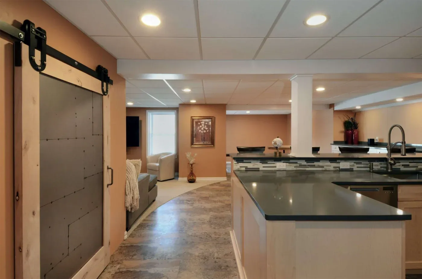 Modern basement with an open layout featuring a kitchen island, sleek lighting, and a sliding barn door. Neutral tones create a warm, inviting ambiance.