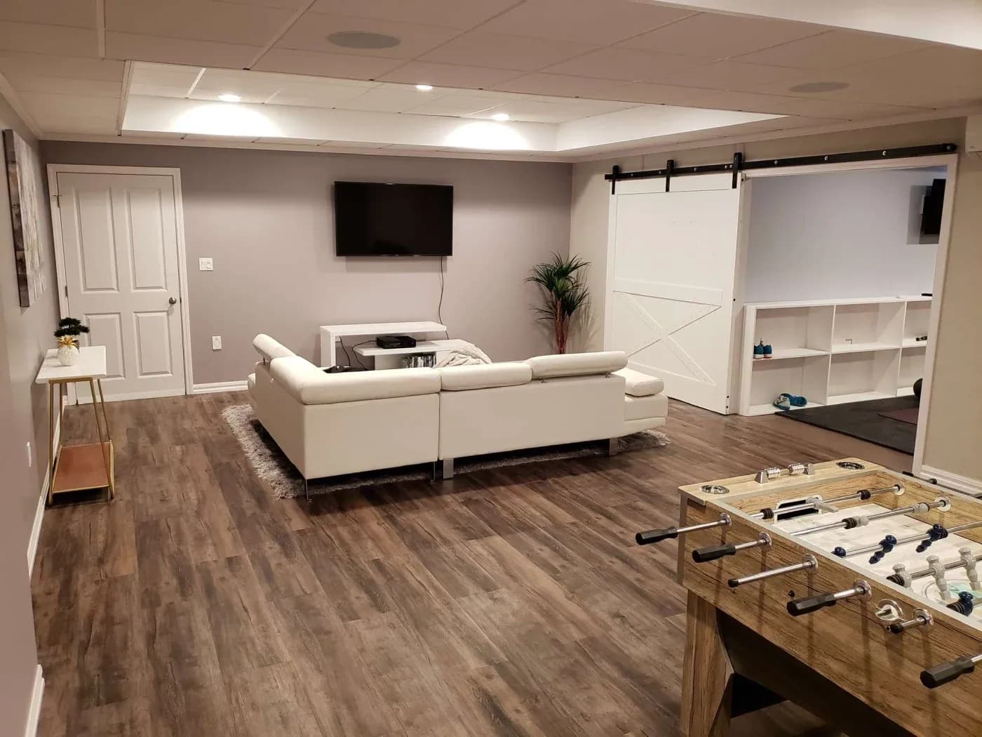 A modern basement with light gray walls and wood flooring features a white sectional sofa facing a wall-mounted TV. A foosball table and barn-style sliding door add to the relaxed vibe.