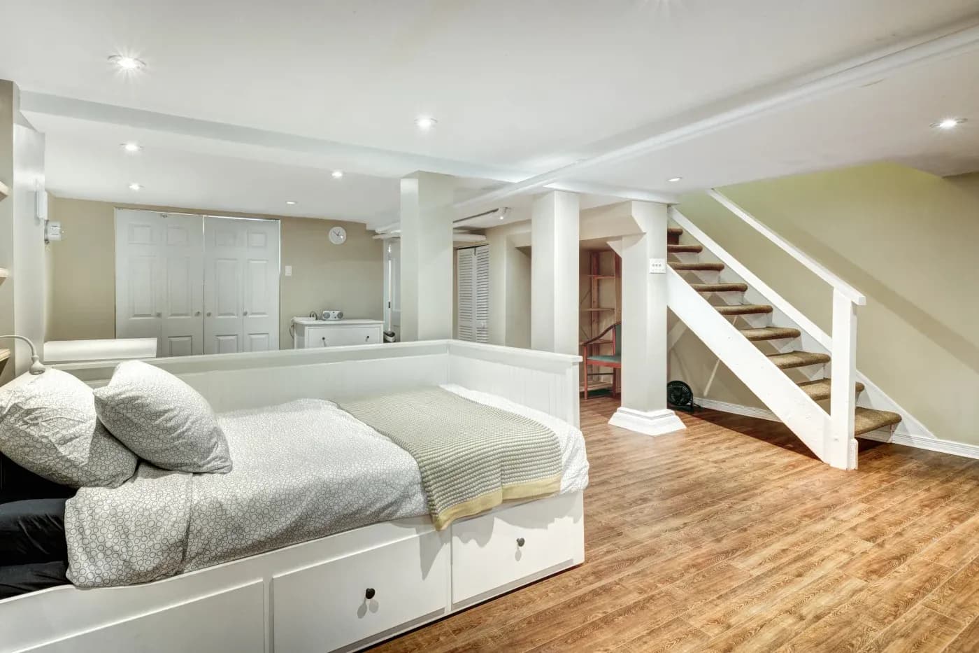 Cozy basement room with wooden floors, featuring a white daybed with gray bedding, a staircase, and storage cabinets. Warm ambient lighting creates a relaxed atmosphere.