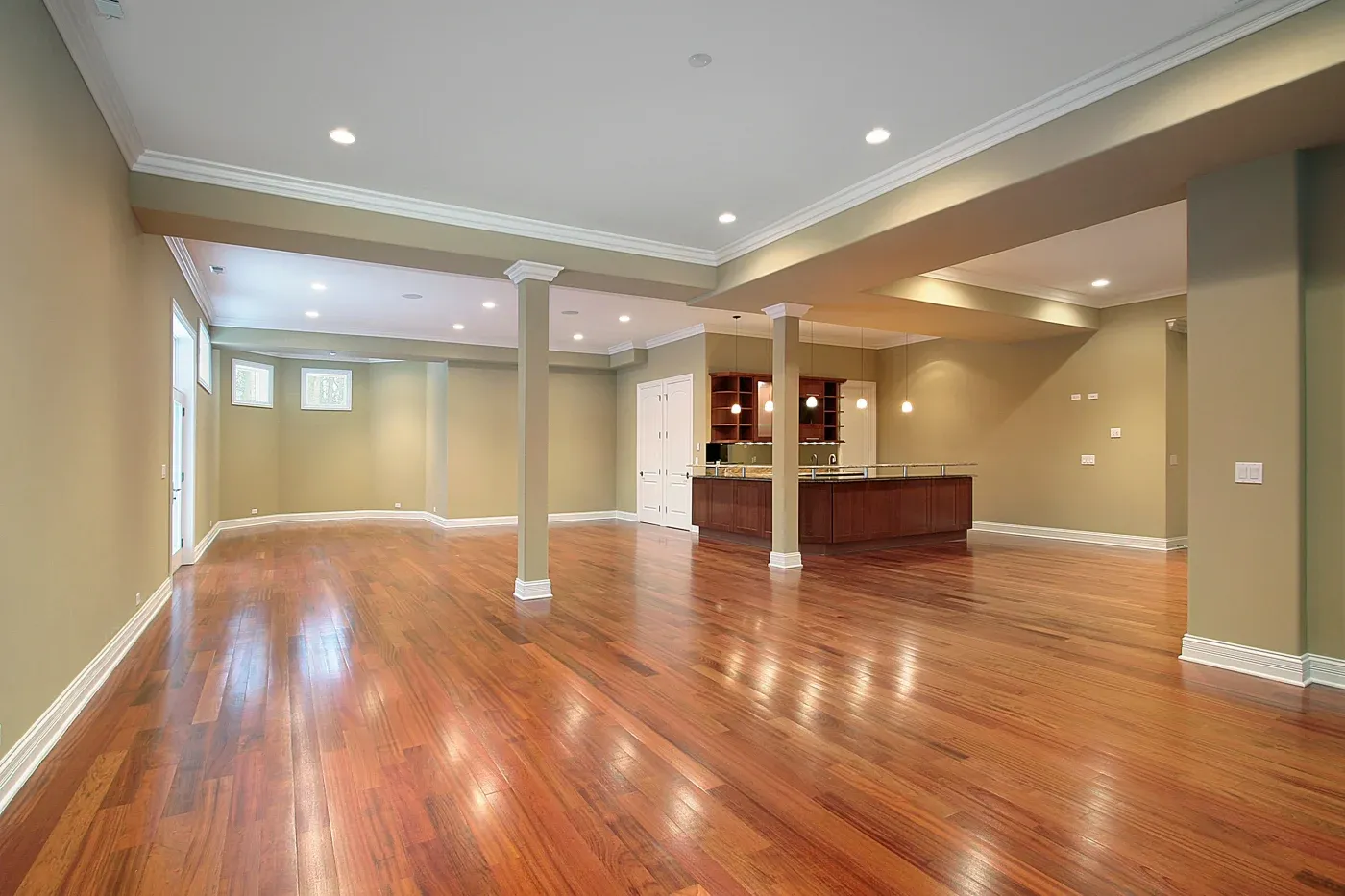 Spacious basement with polished wooden floors, beige walls, and recessed lighting. A cozy kitchenette with dark wood cabinets is at the far end.