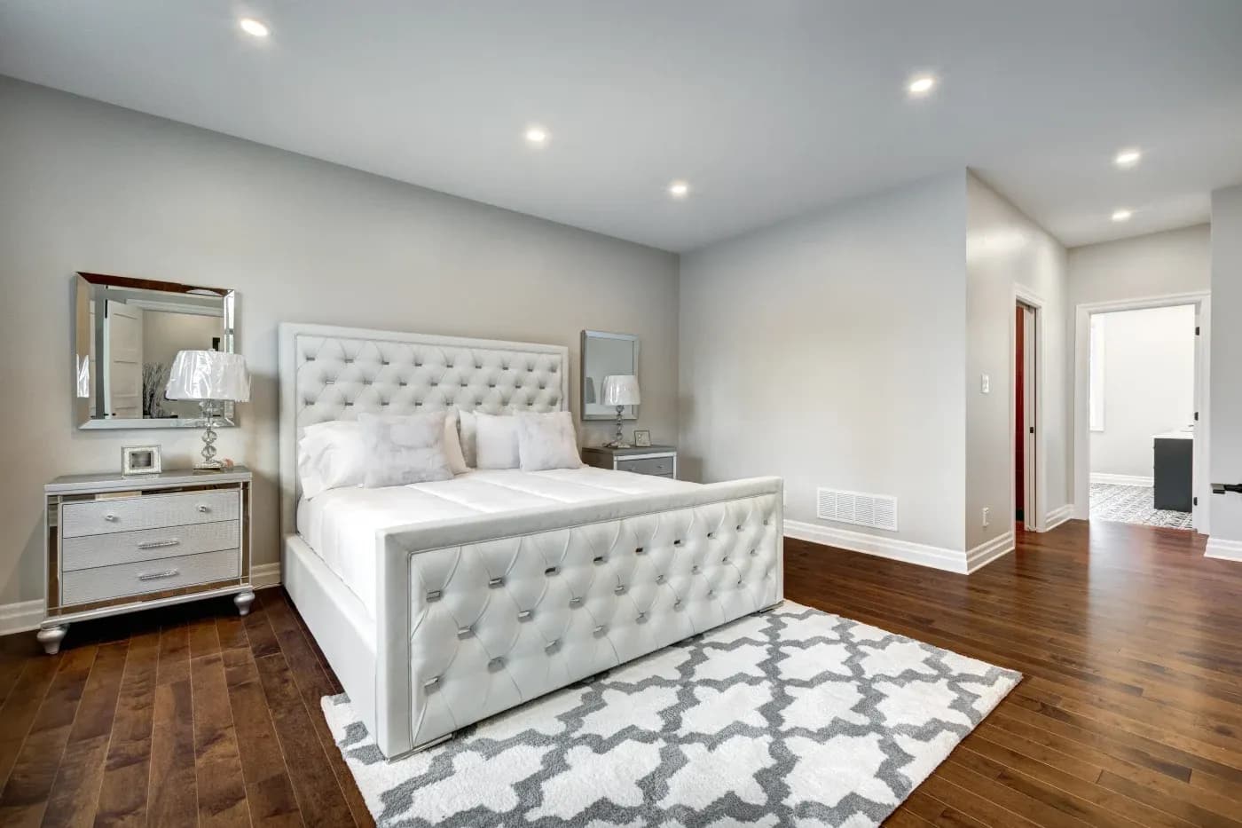 Elegant bedroom with a tufted white bed, two mirrored nightstands with lamps, and a geometric rug on dark wood flooring. Soft, modern ambiance.