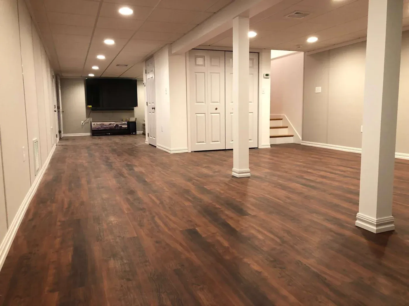 Spacious modern basement with polished wood floors and cream walls, featuring ceiling lights, columns, and a TV on a stand, conveying a cozy atmosphere.