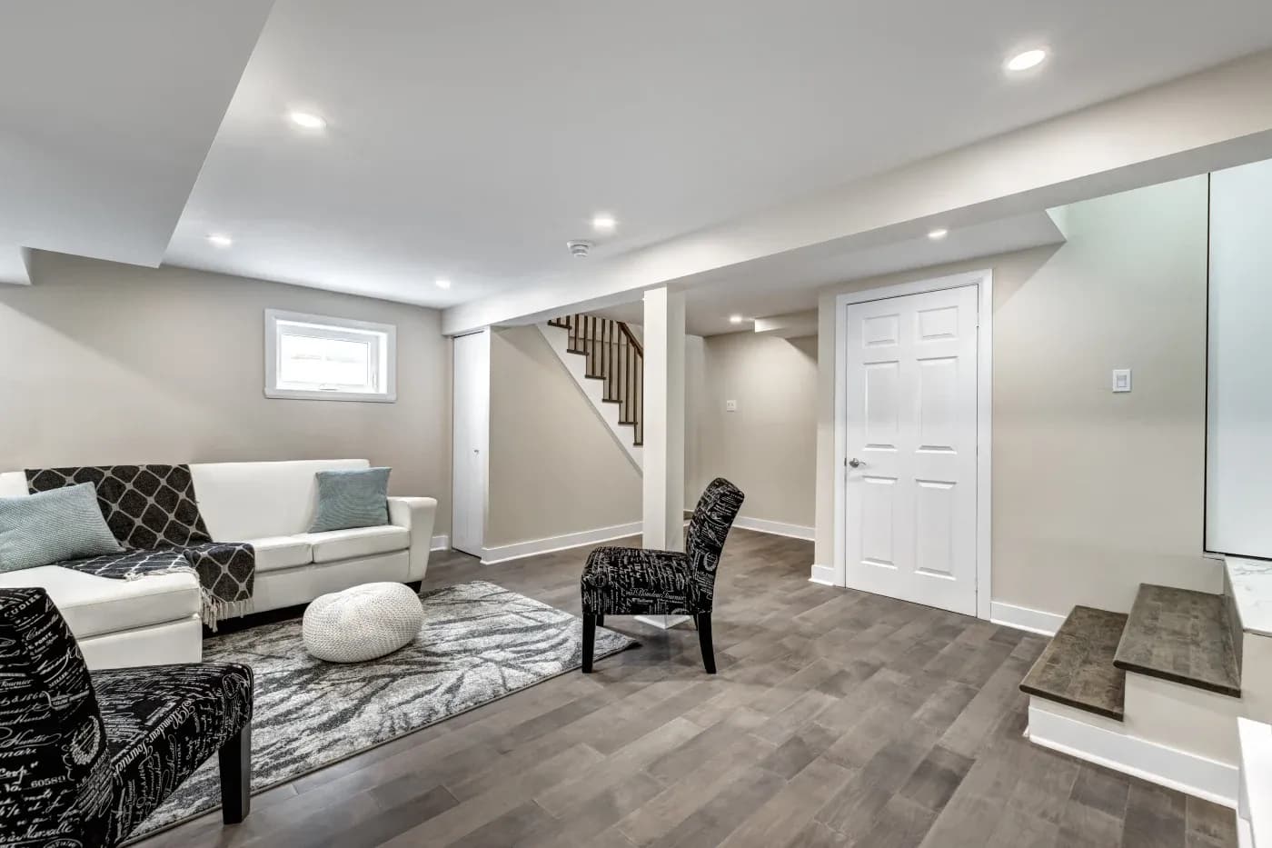 Modern basement living room with beige walls and dark wood flooring. Features a white sofa with pillows, patterned chairs, a rug, and stairs. Bright, cozy ambiance.