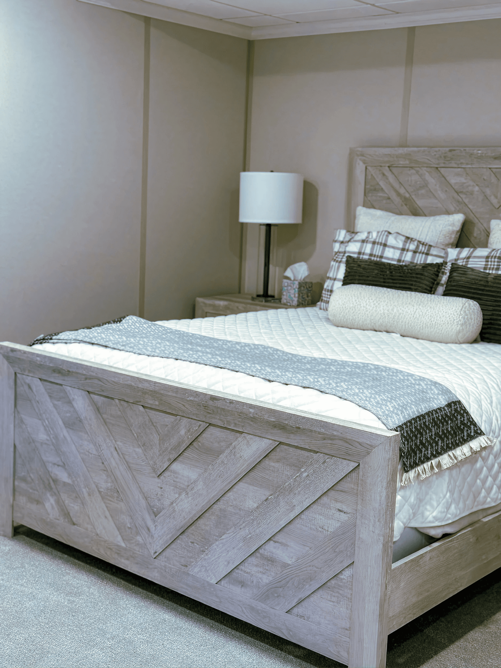 A cozy bedroom with a rustic wooden bed featuring geometric patterns. The bed is dressed in white and plaid linens with decorative pillows. A lamp and tissue box are on the nightstand, creating a serene, inviting atmosphere.