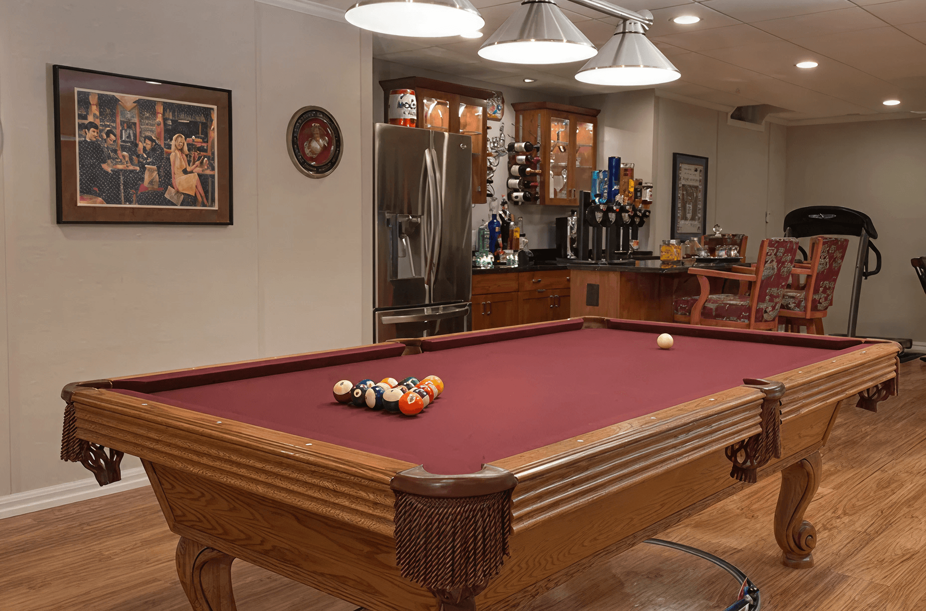 Cozy basement with a red pool table centered, surrounded by bar area, stools, and dim lighting. Art decorates the walls, creating a relaxed atmosphere.