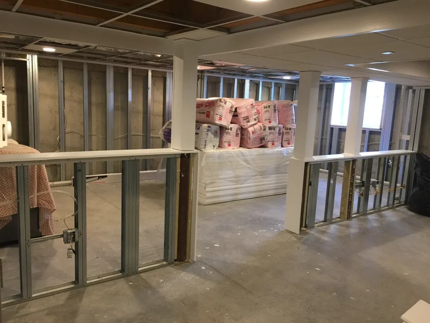 A partially finished basement features exposed metal framing and insulation materials stacked in the center. The space is lit with overhead lighting, creating a work-in-progress feel.