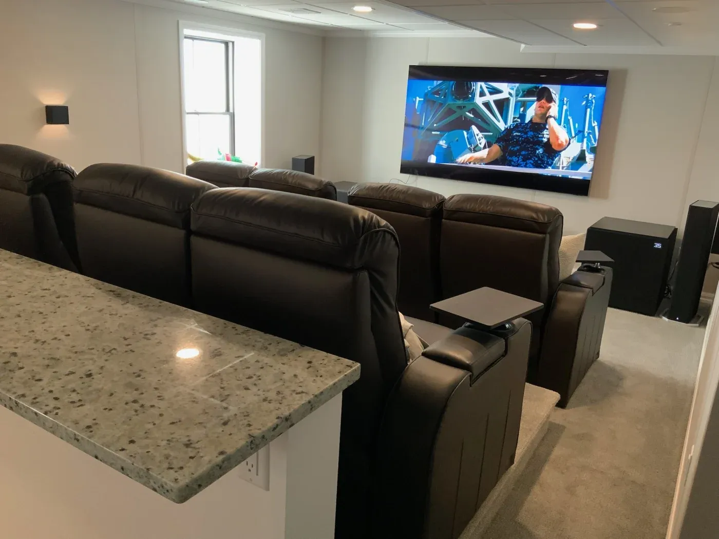Home theater with three rows of black leather recliners facing a large screen showing an action movie. A granite counter is in the foreground. Cozy atmosphere.