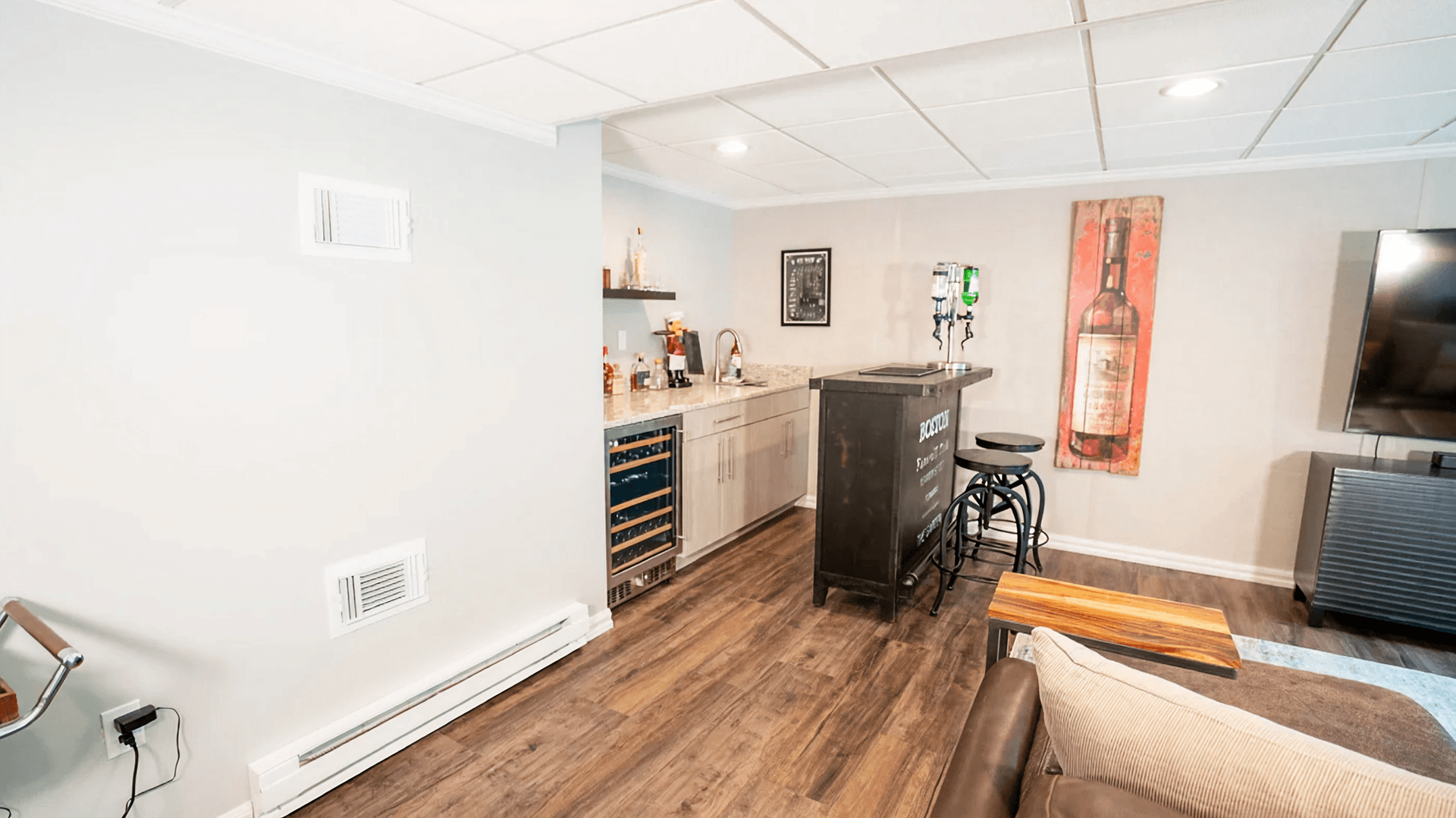 A modern basement bar with light gray walls and a wood floor. It features a countertop with liquor bottles, bar stools, and a beverage cooler. A large wine bottle painting adds a cozy touch.