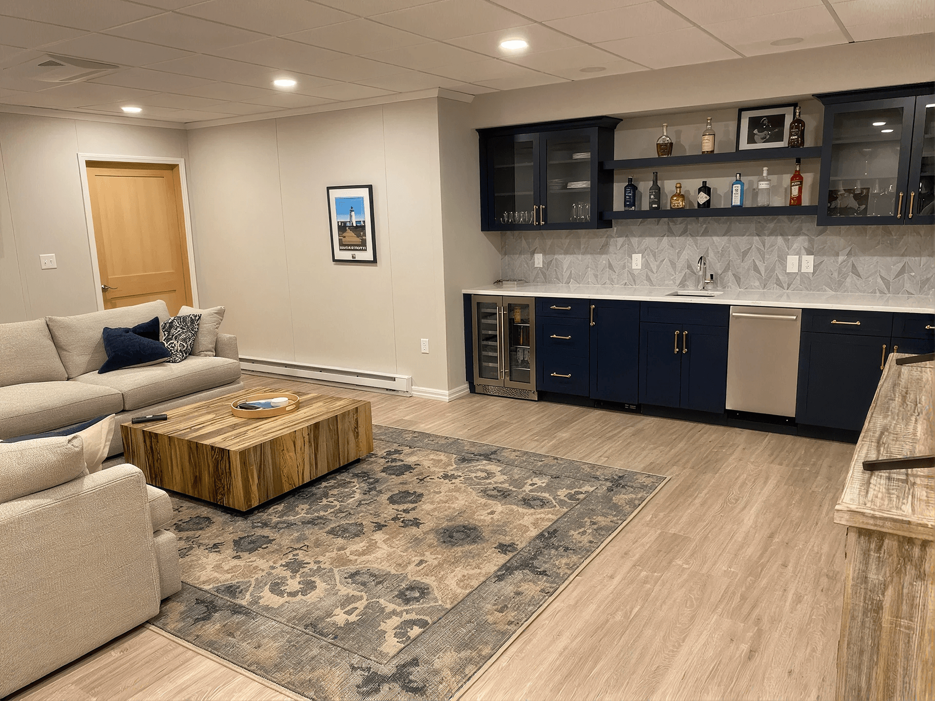 Stylish basement lounge with a gray sectional, wooden coffee table, and patterned rug. Navy cabinets and bar with bottles and glassware evoke a cozy, modern feel.