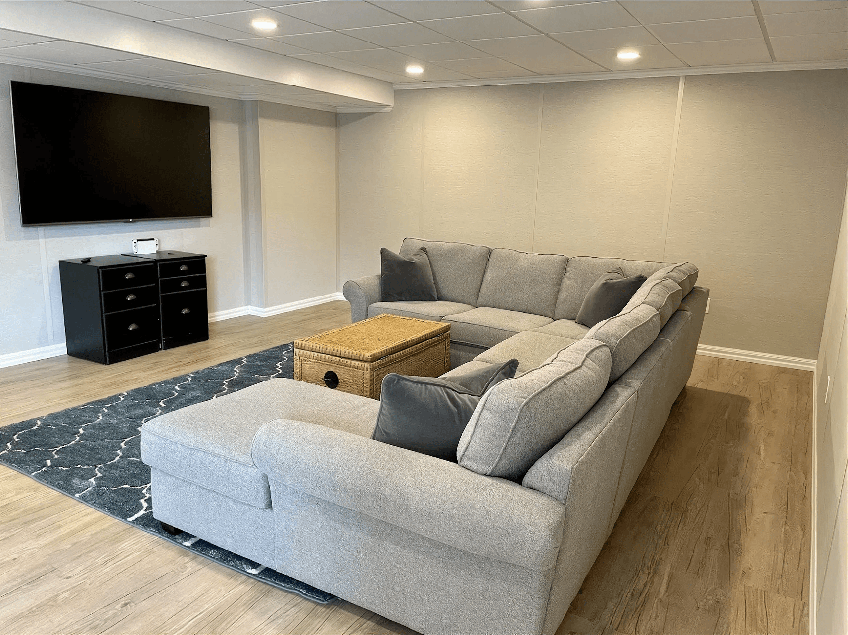 A cozy living room features a large gray sectional sofa and a wicker coffee table on a patterned blue rug. A flat-screen TV is mounted on the wall.