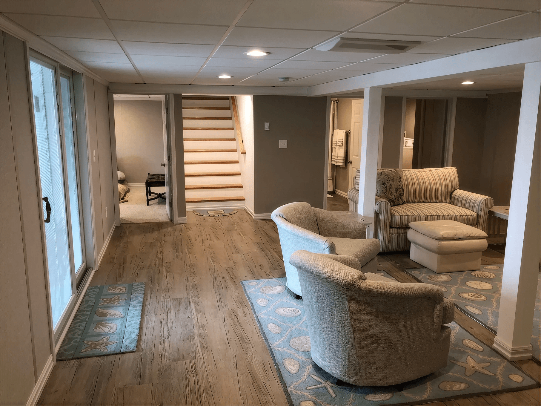 Spacious basement with wooden flooring features two beige armchairs and a striped couch on blue patterned rugs. Stairs and sliding glass doors visible.