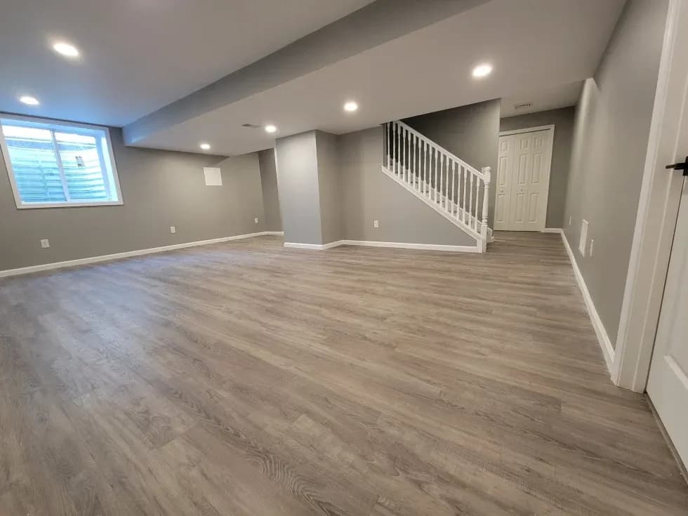 A spacious, modern basement with gray walls and wood flooring, featuring a window, staircase with white railing, and recessed lighting, creating a cozy atmosphere.